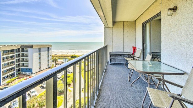 Un balcon avec une table et des chaises en verre, donnant sur une plage et un parking.