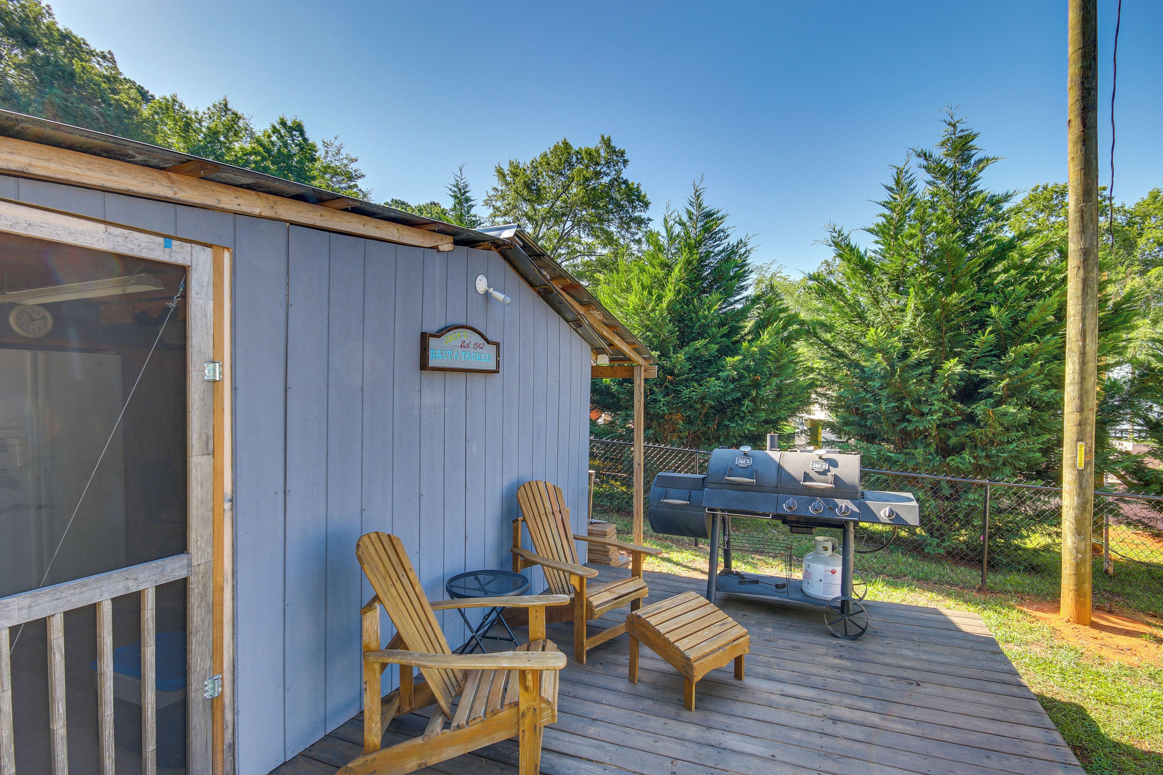 A wooden deck with Adirondack chairs and a grill, a covered area with a sign, and a forested background.