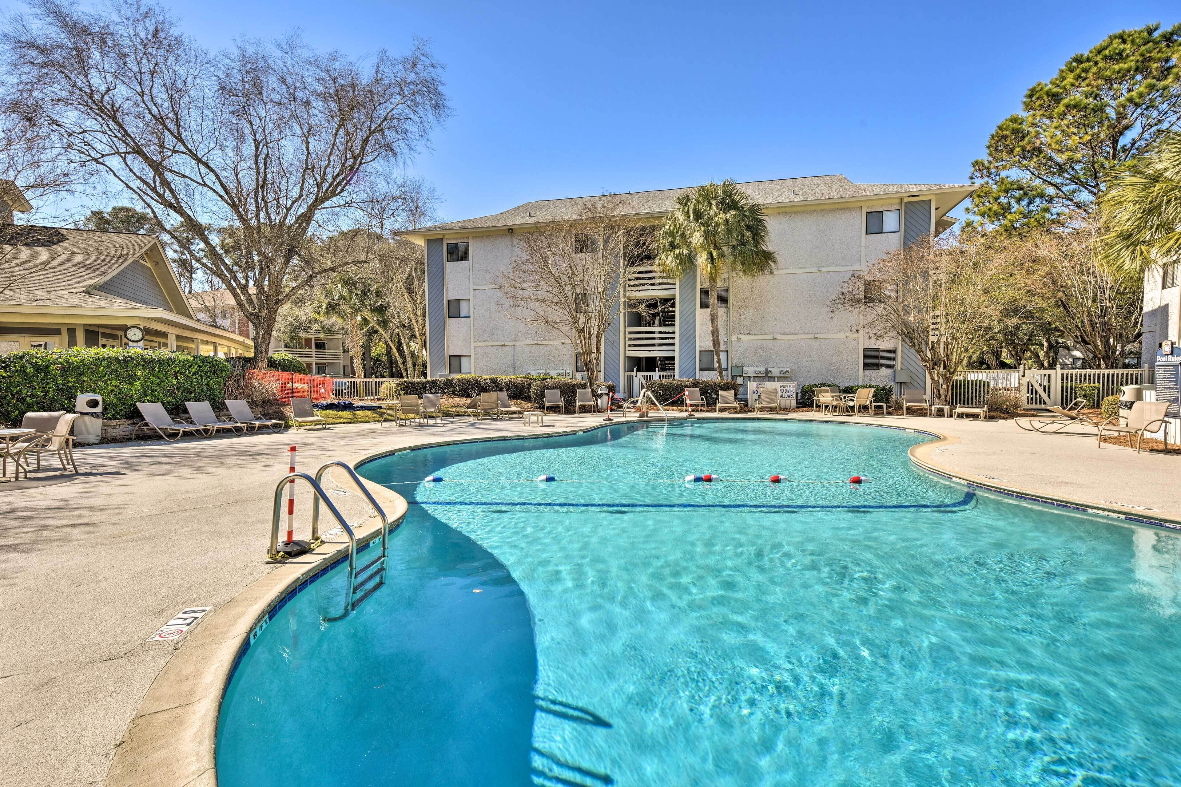 A clear swimming pool with a diving board, surrounded by lounge chairs and a building in the background.