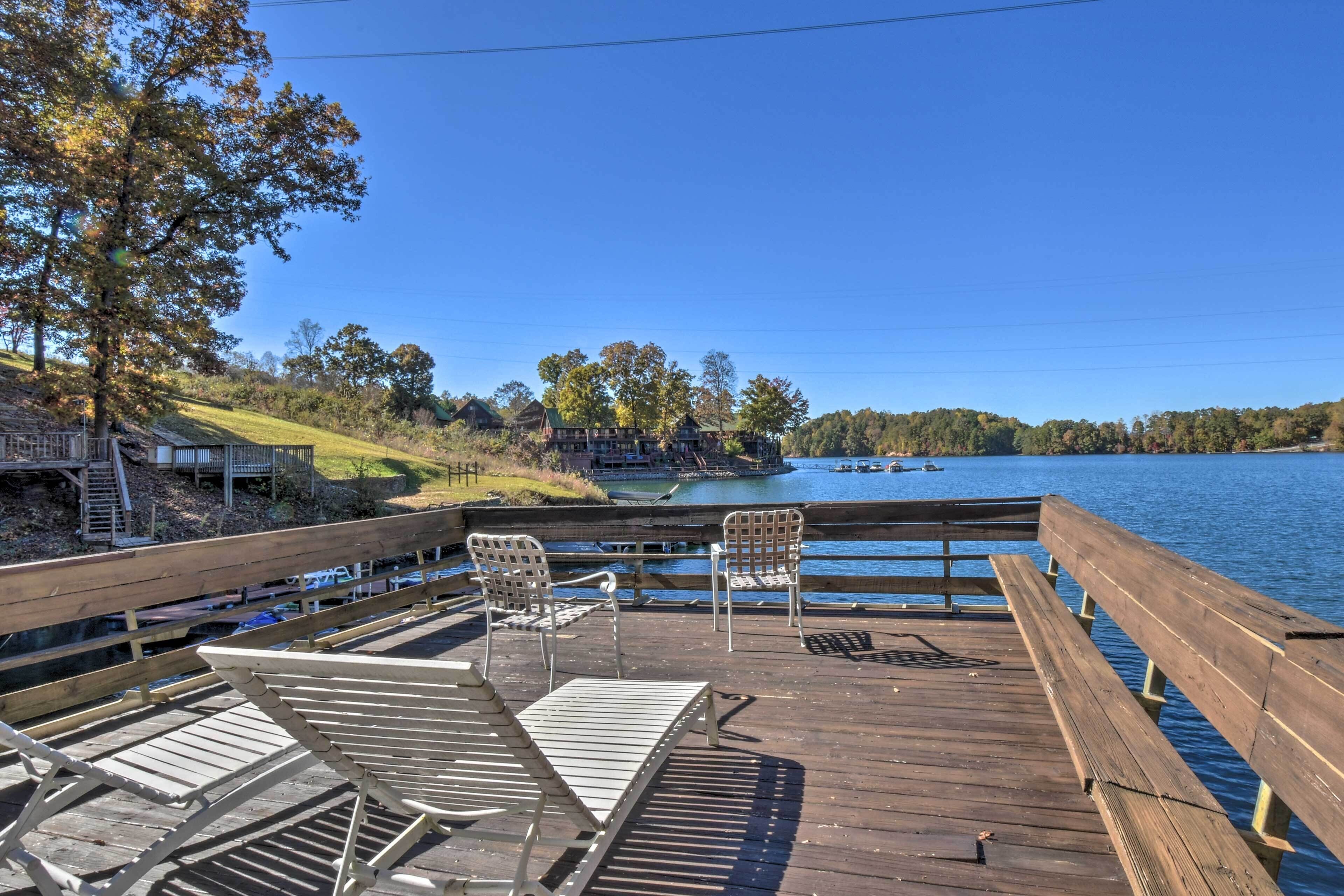A wooden deck with lounge chairs overlooking a lake, surrounded by trees and a distant building.