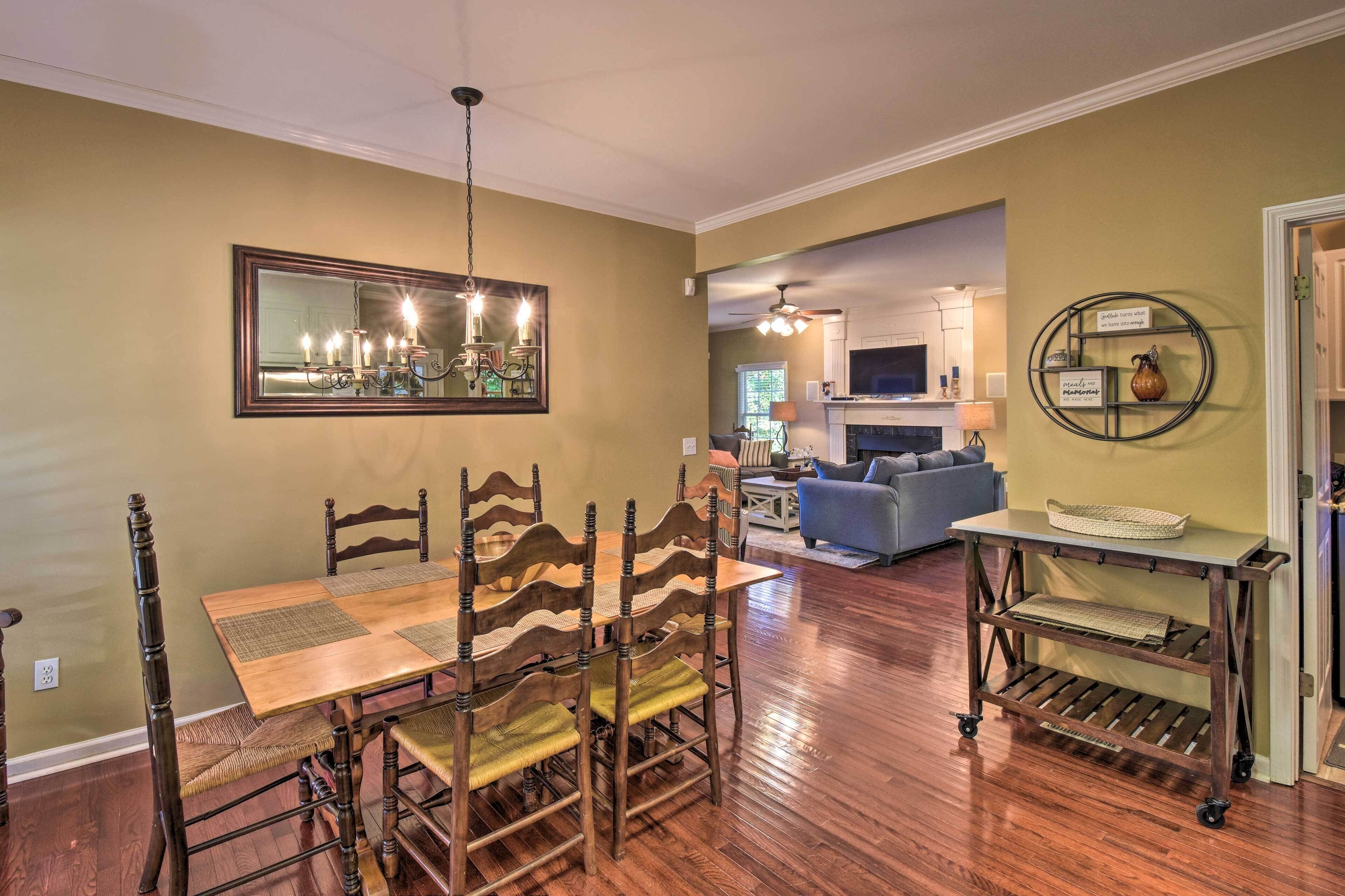 A dining area with a wooden table and chairs, a chandelier, and a kitchen in the background.