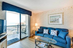 A living room with a blue sofa, a glass coffee table, a television, and a balcony with outdoor furniture.