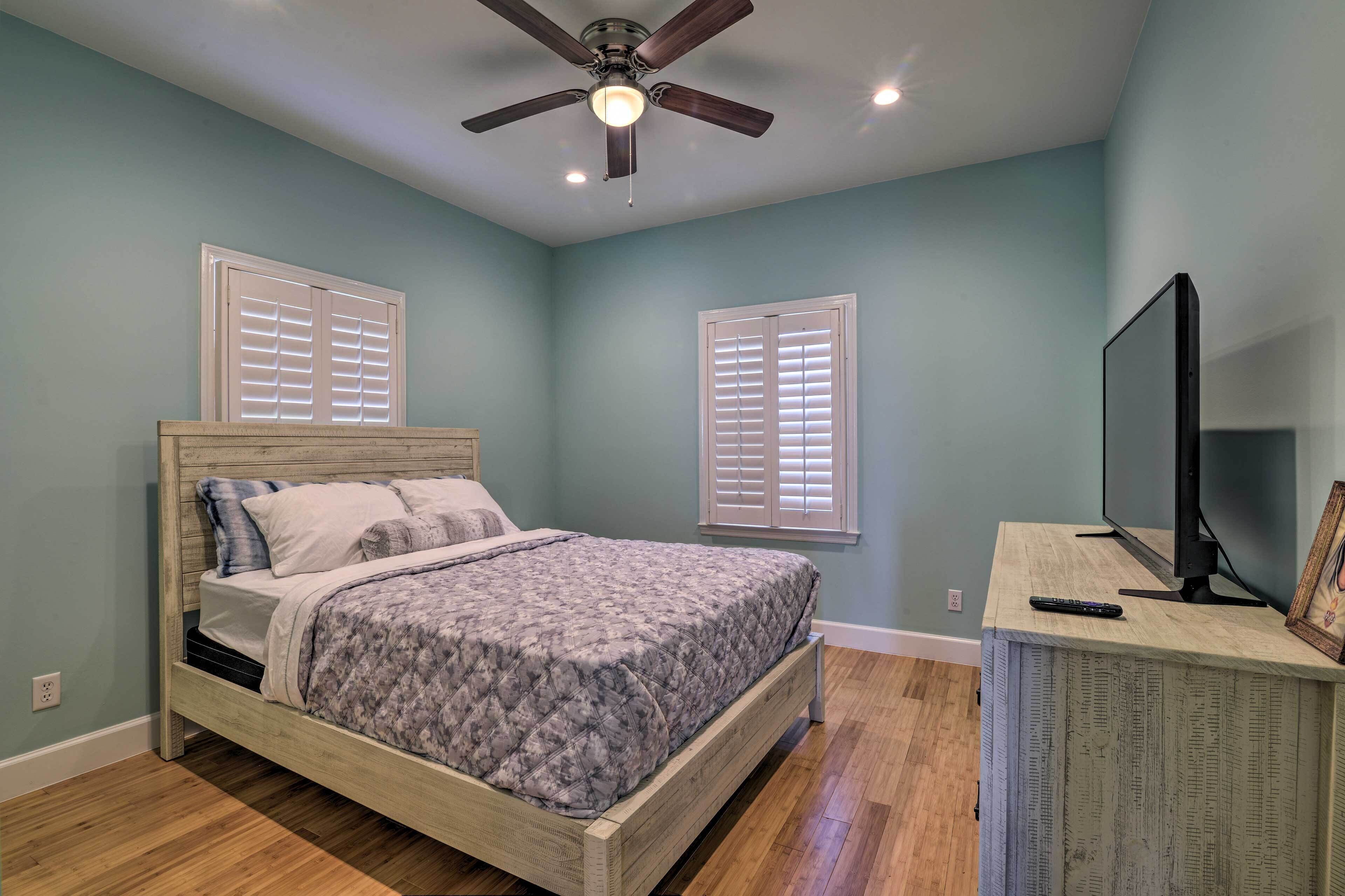 A bedroom with a bed, a ceiling fan, a TV, and two windows with blinds.