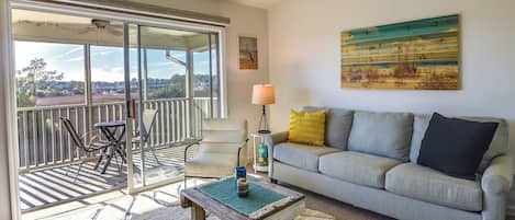 A living room with a grey sofa, a coffee table, a lamp, and a view through a sliding glass door to the outside.