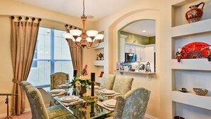 A dining area with a glass table, four upholstered chairs, a chandelier, and a view into the kitchen.