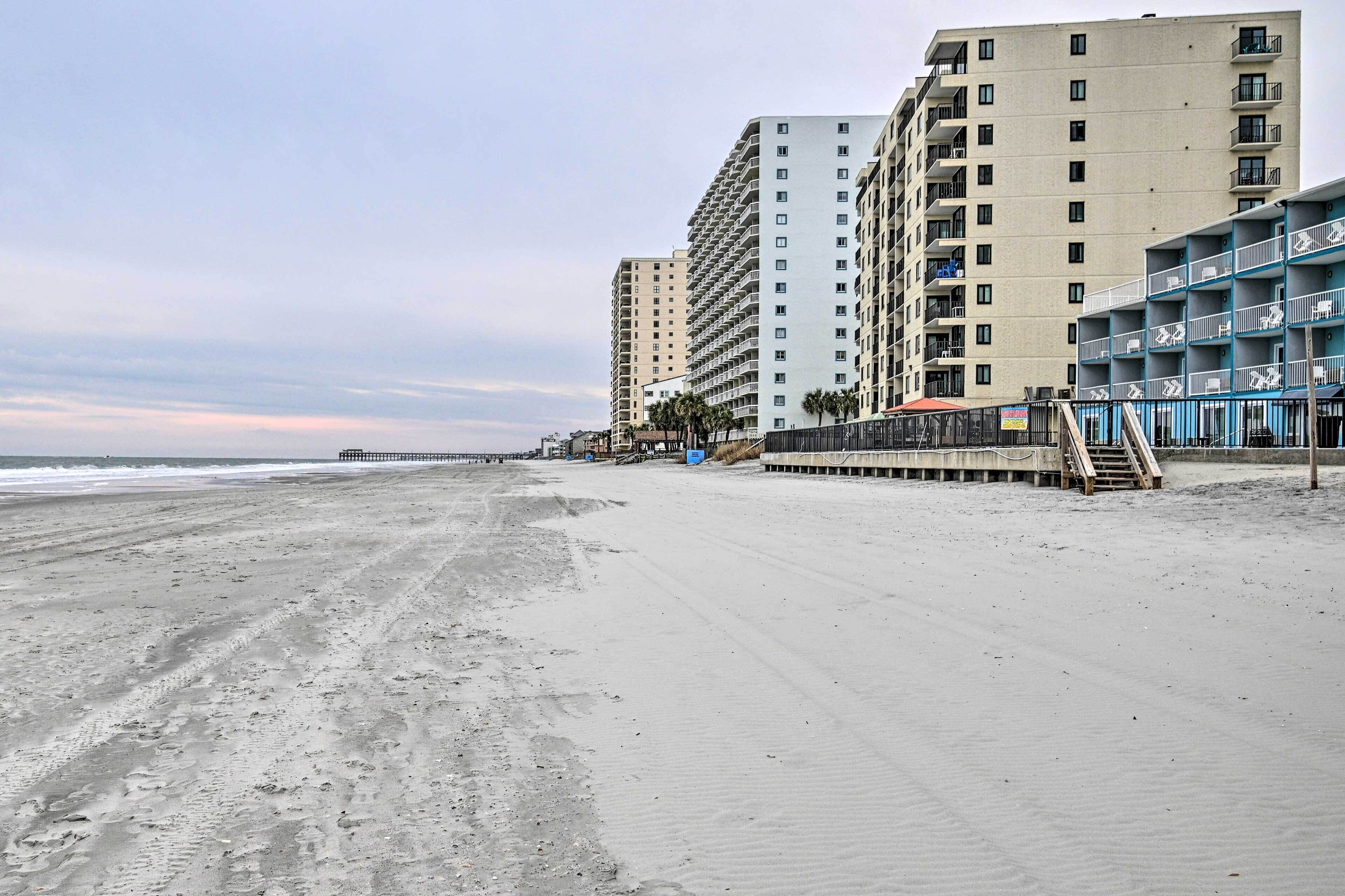 A beachside hotel with multiple floors, a boardwalk, and a pier in the background.