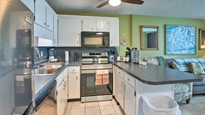 Cocina compacta con armarios blancos, microondas, horno y lavavajillas. Cuenta con ventilador de techo, lavabo y basurero. Las paredes son verdes y hay un cuadro en la pared.