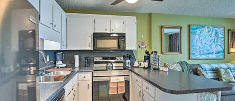 A compact kitchen with white cabinets, a microwave, an oven, and a dishwasher. There is a ceiling fan, a sink, and a trash can. The room has green walls and a painting on the wall.