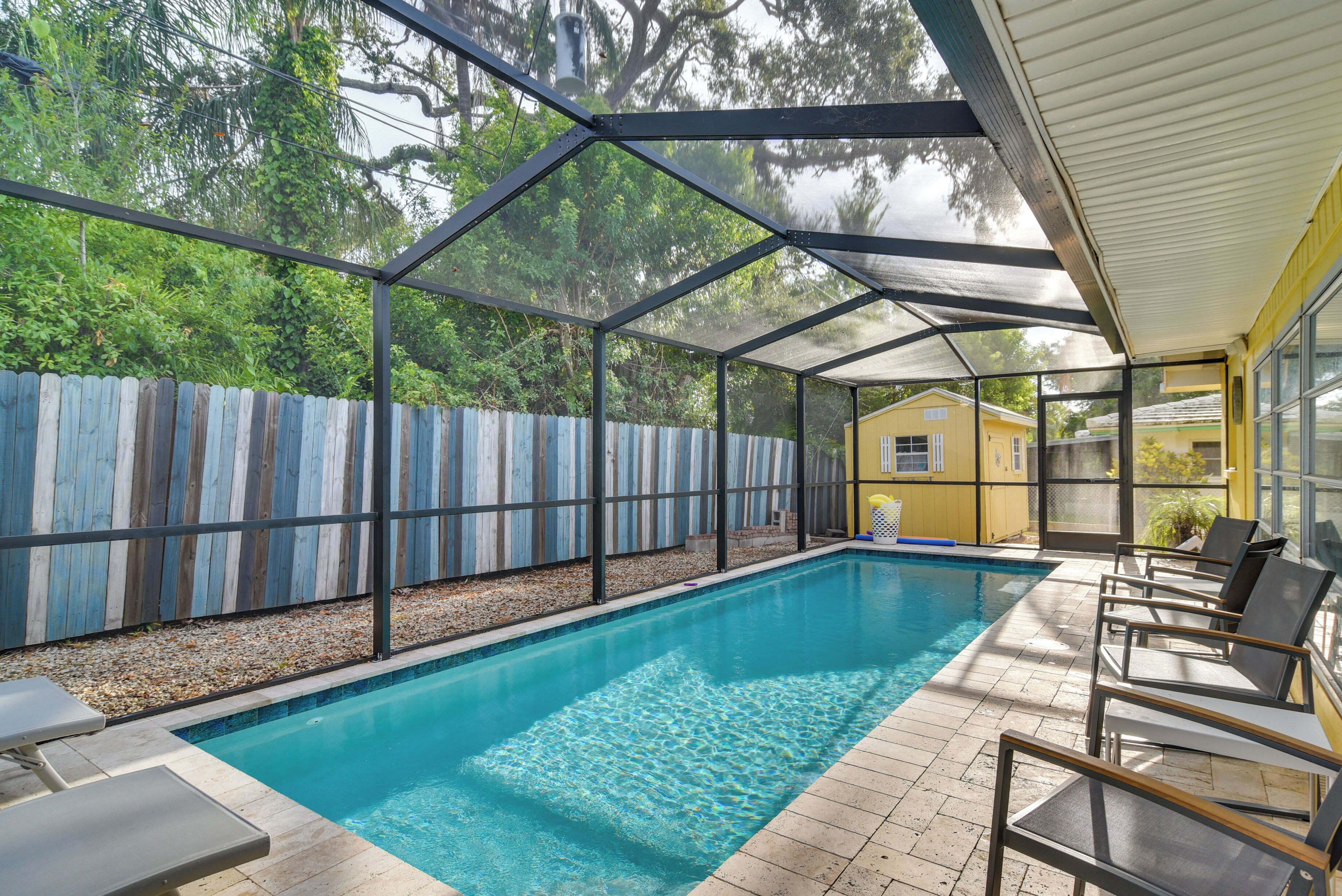 Une piscine couverte, entourée d’une clôture en bois, avec des chaises longues et un bâtiment jaune en arrière-plan.
