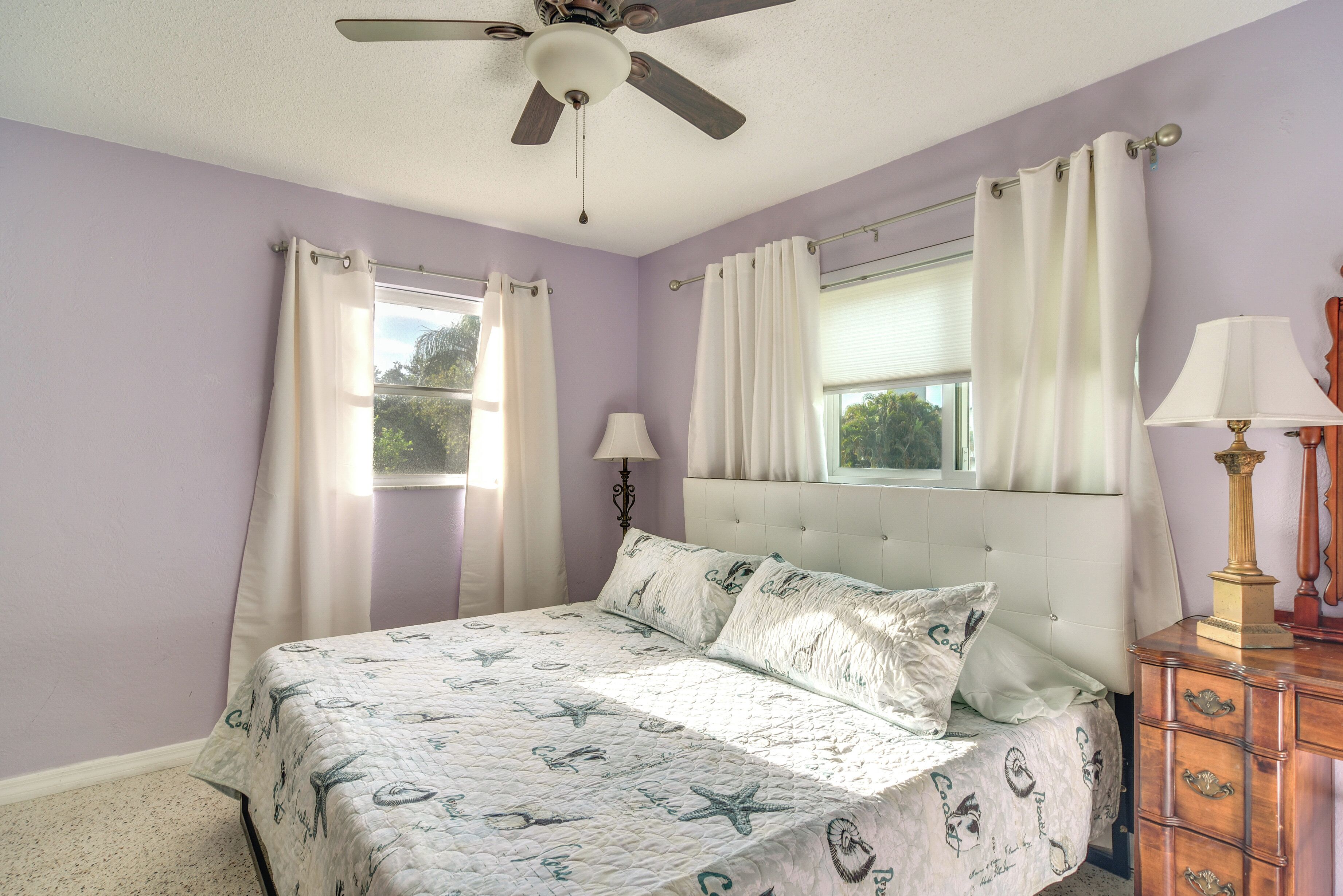 Une chambre à coucher comprenant un lit, un ventilateur de plafond, une commode et deux fenêtres avec des rideaux.