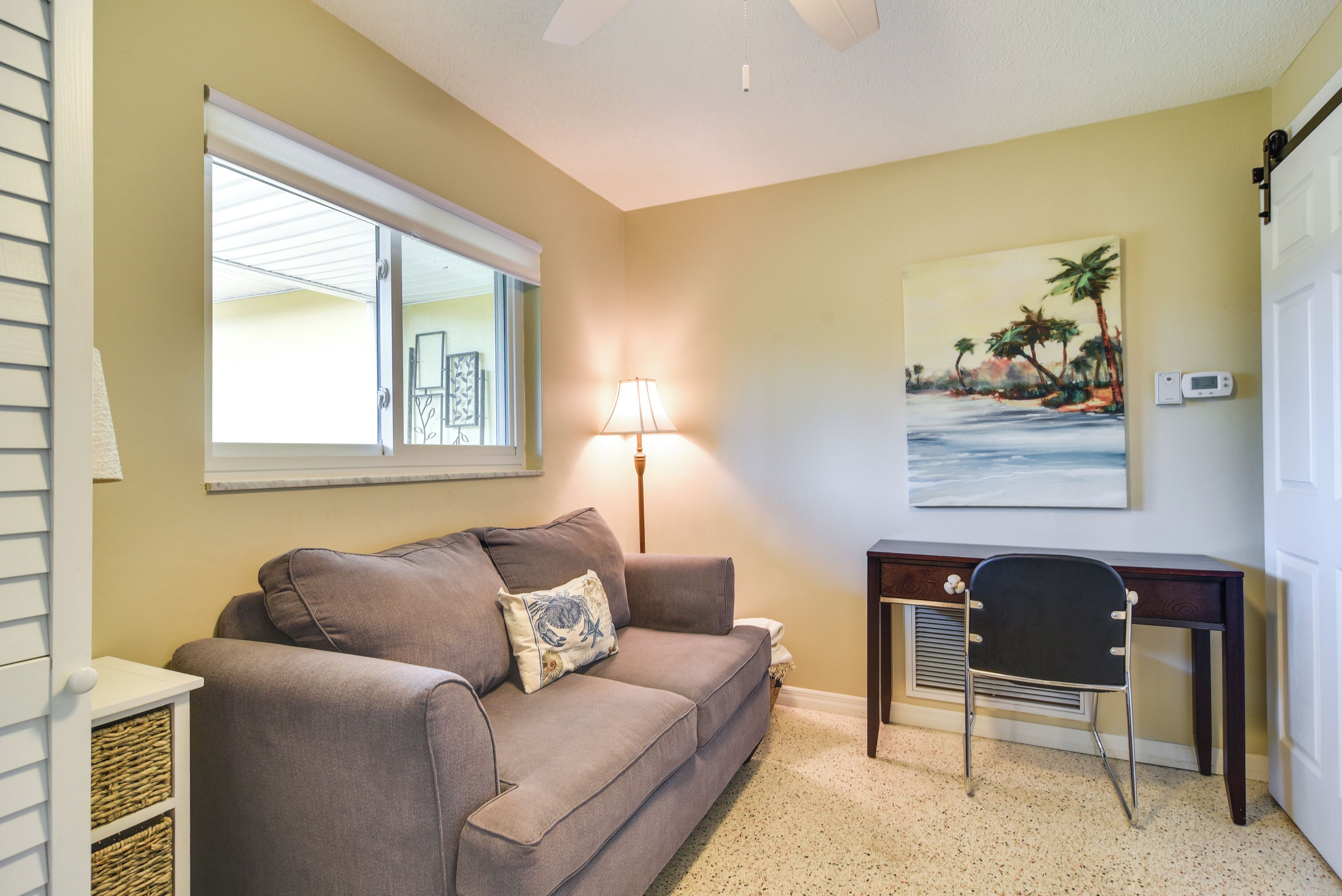 A living room with a beige wall, a brown sofa, a lamp, a desk, and a painting of a beach scene.