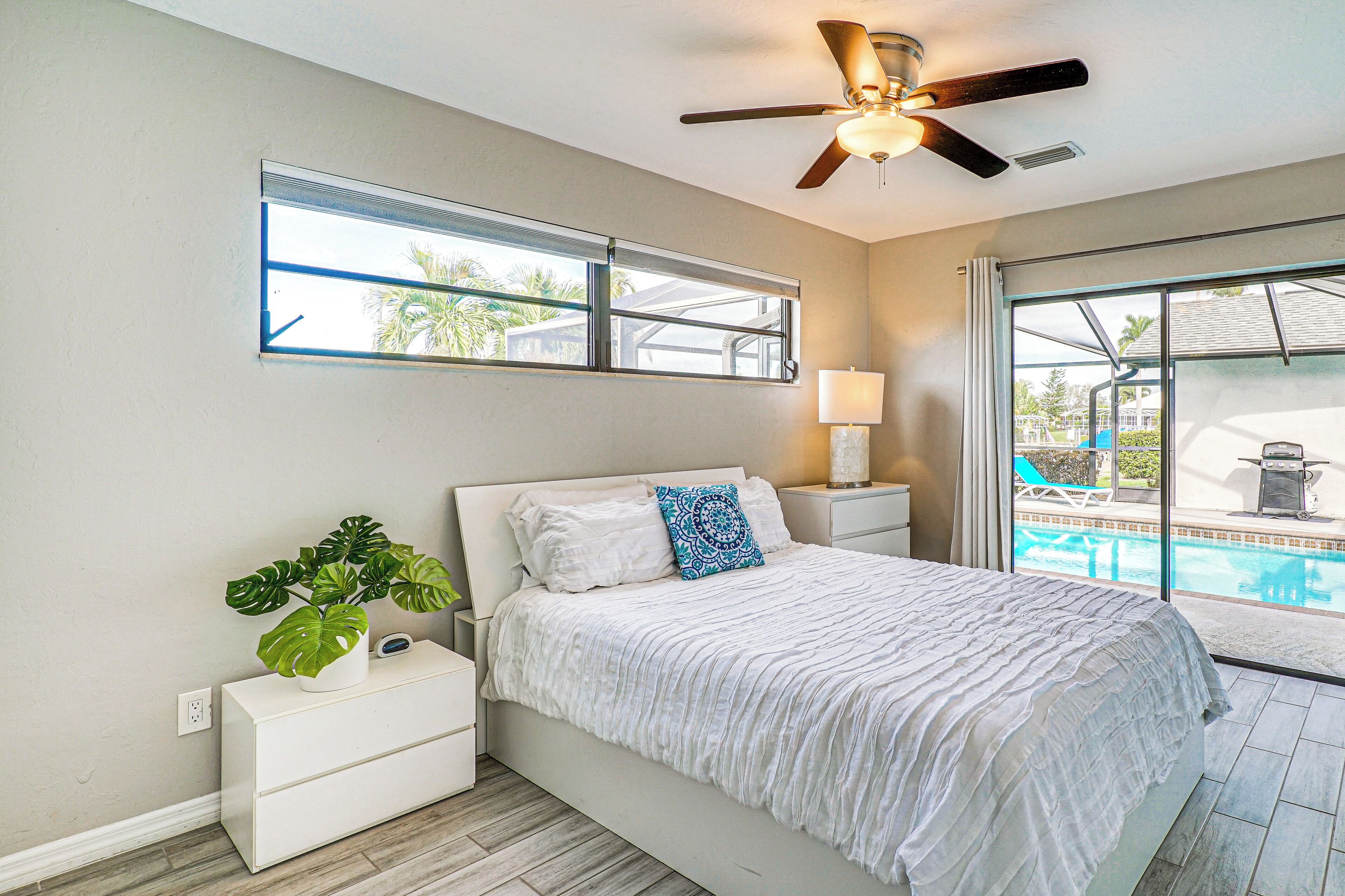 A bedroom with a bed, a nightstand, a bedside lamp, a ceiling fan, a window, and a view of a swimming pool.