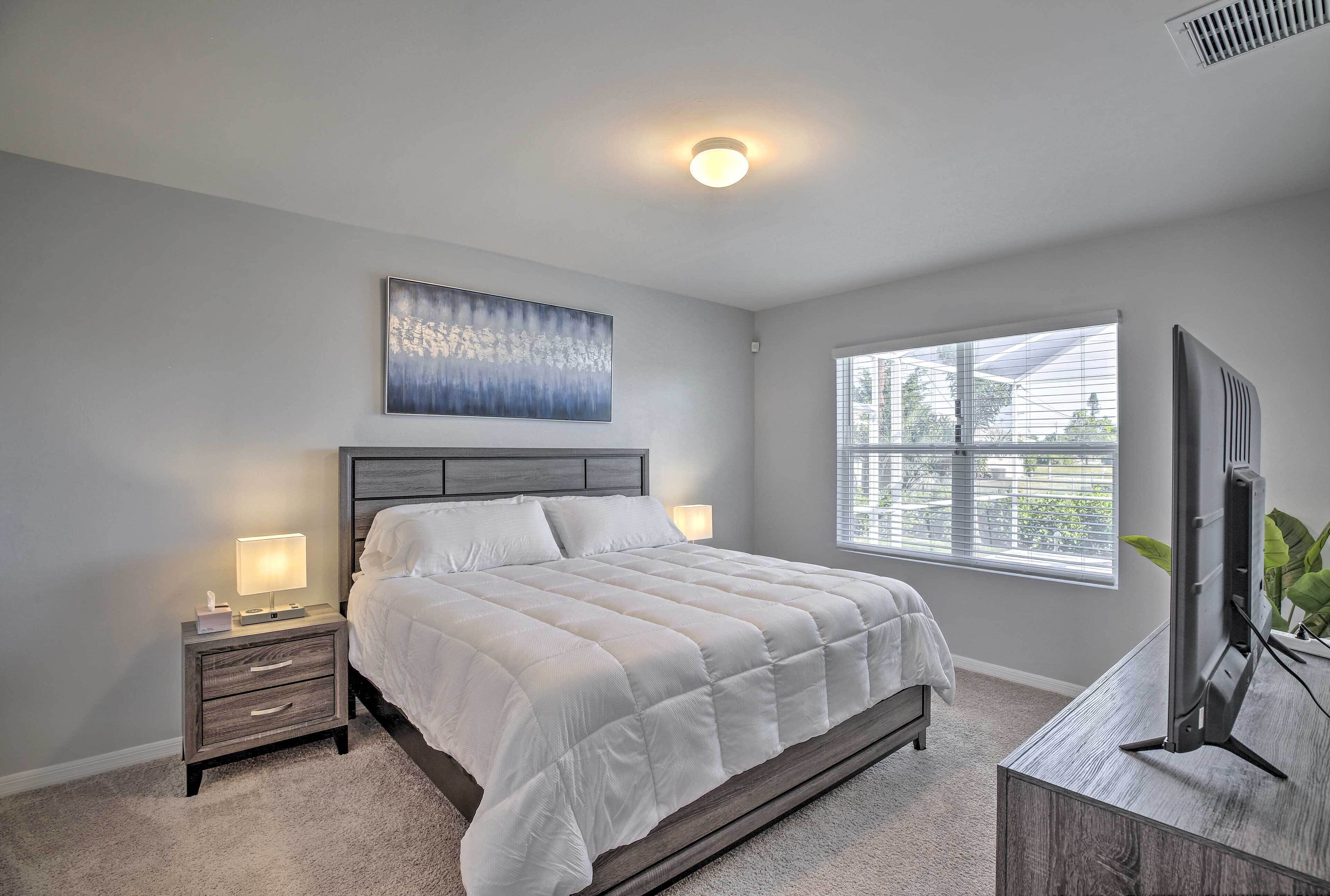 A bedroom with a bed, nightstand, lamp, artwork, window, and TV stand.