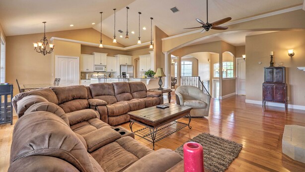 A spacious living room with a large sectional sofa, a coffee table, and a red vase. There are ceiling fans, pendant lights, and a view into the kitchen area.