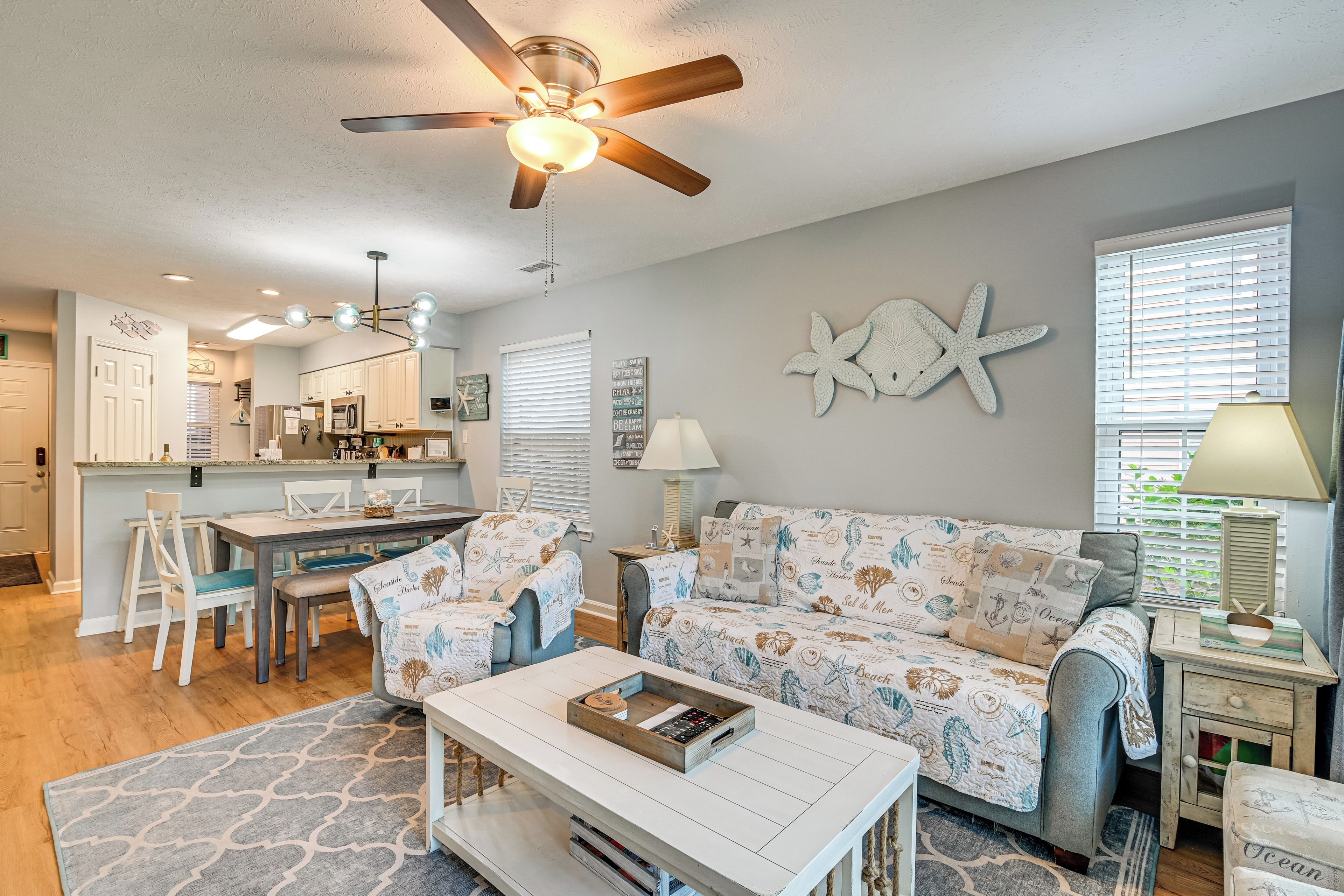 A spacious living room with a sofa, coffee table, and dining area. A ceiling fan and kitchen are visible in the background.