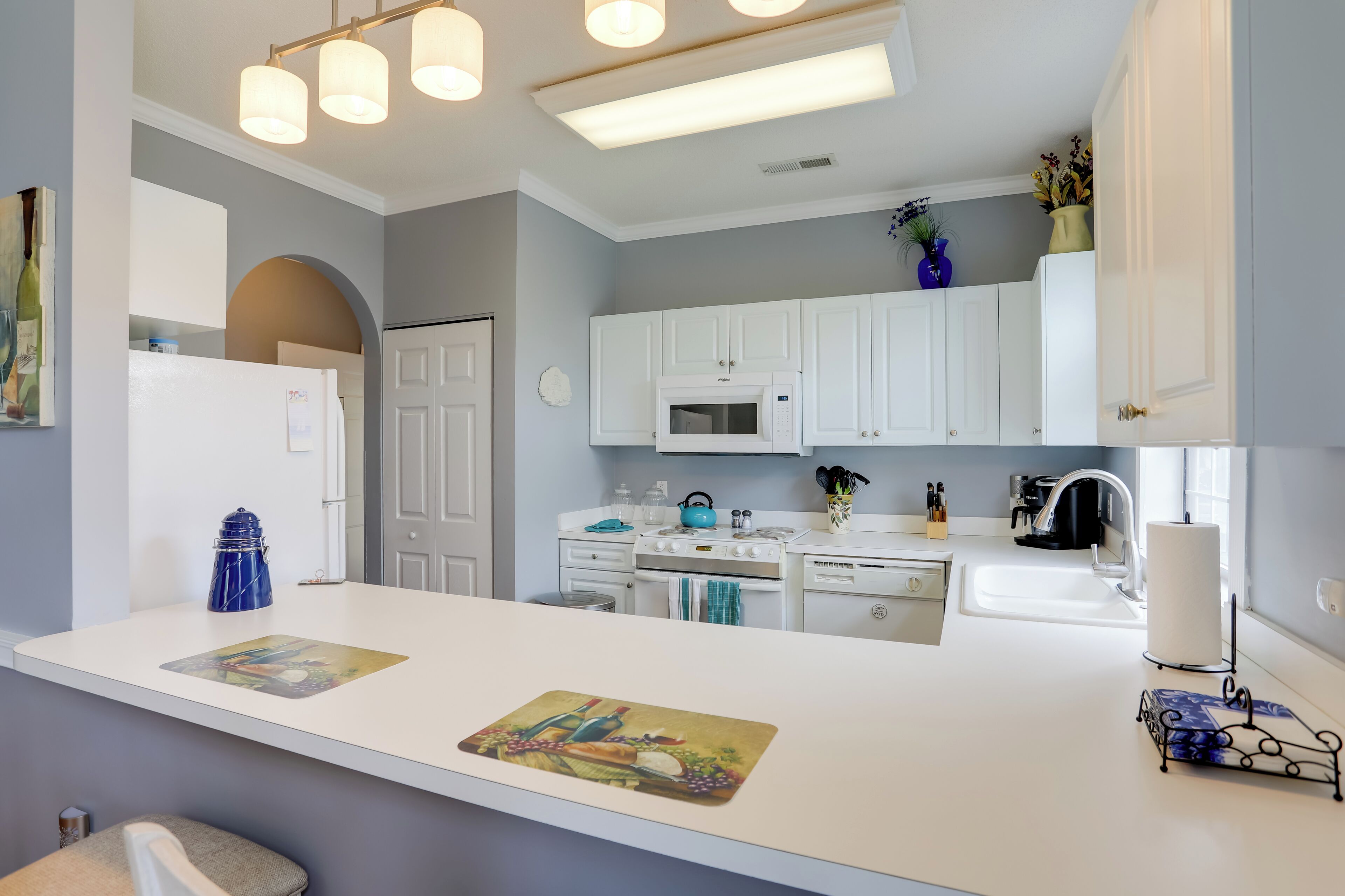 Una cocina moderna con armarios blancos, un microondas, una estufa y un lavabo. Hay una tetera azul y un florero con flores en la encimera.