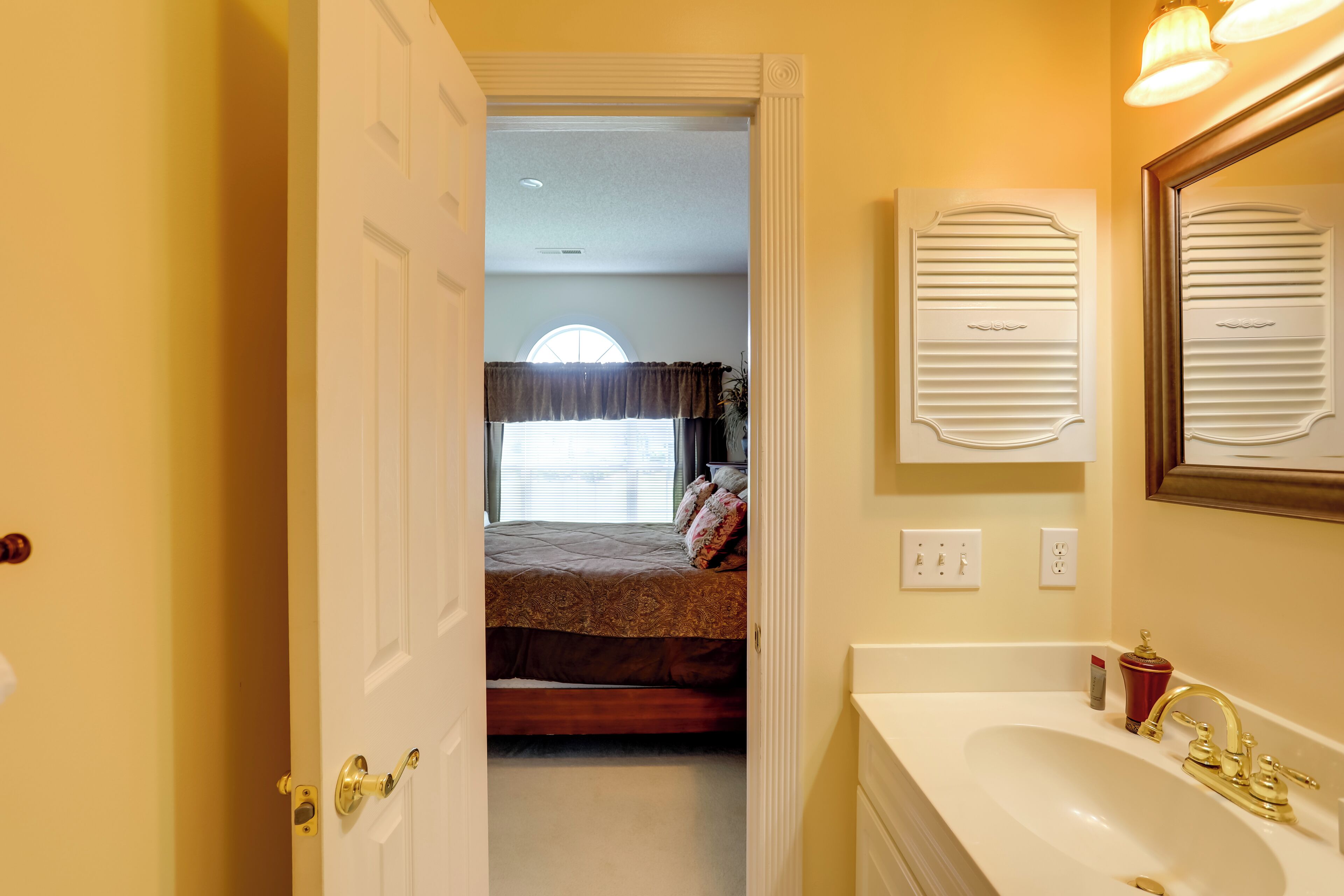 A bedroom with a bed, a window with blinds, a bedside table, and a bathroom with a sink and mirror.