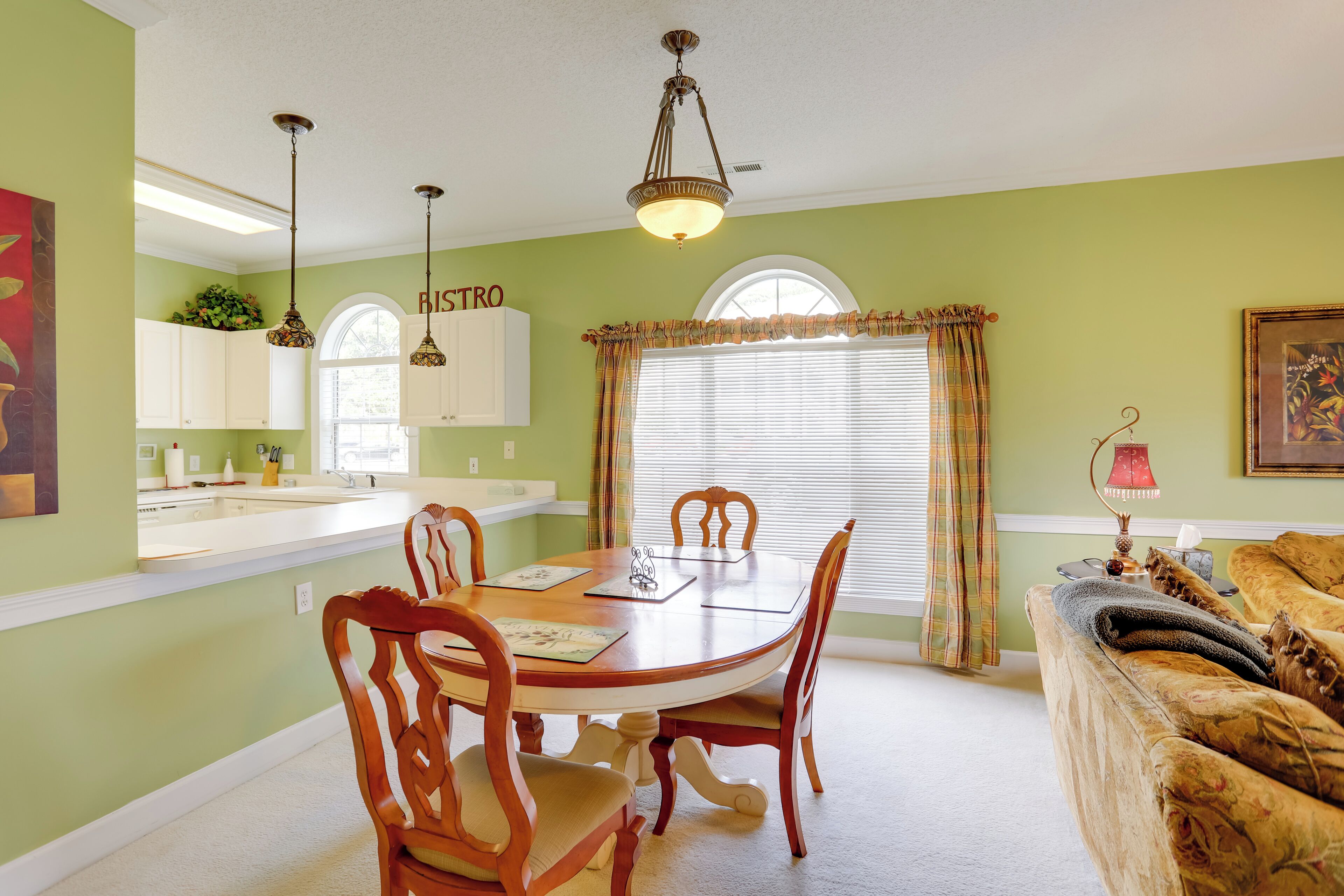 A dining area with a wooden table and chairs, a kitchen with white cabinets, and a window with curtains.