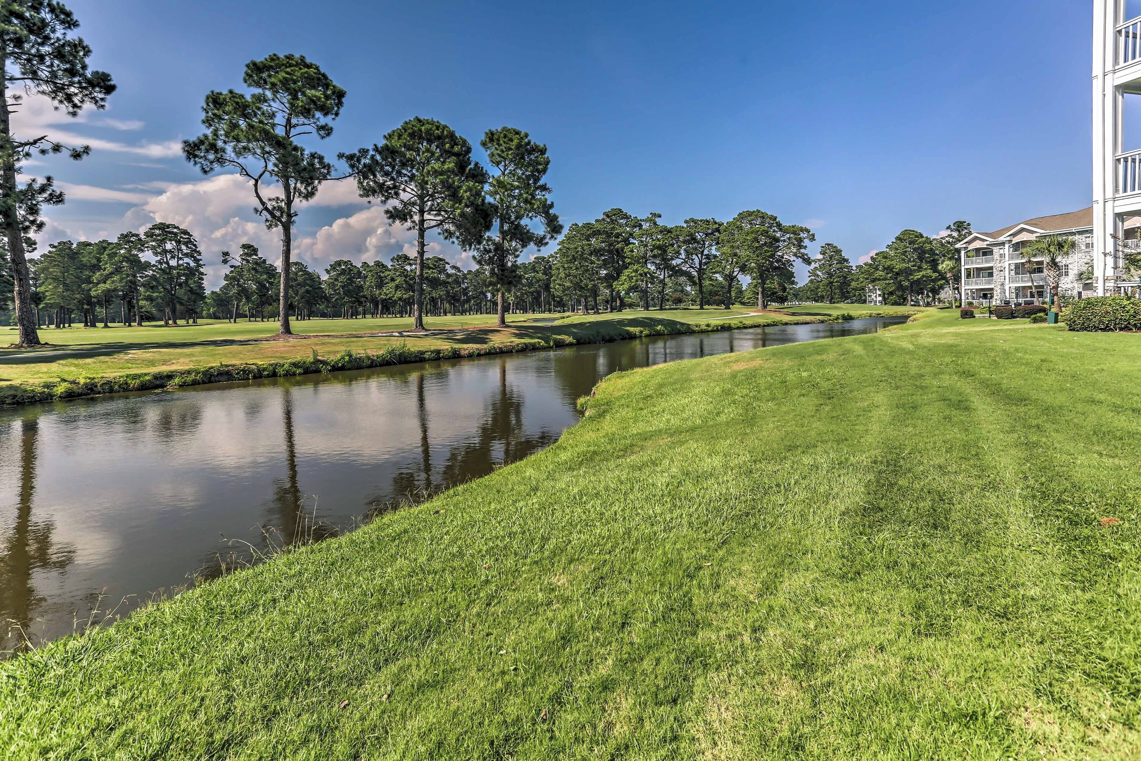 A golf course with a water body, trees, and a building with multiple floors and balconies.