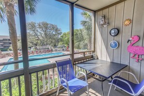 Ein Balkon mit Blick auf einen Swimmingpool, blaue Stühle, ein Tisch und Wanddekoration, darunter ein Flamingo und Uhren.