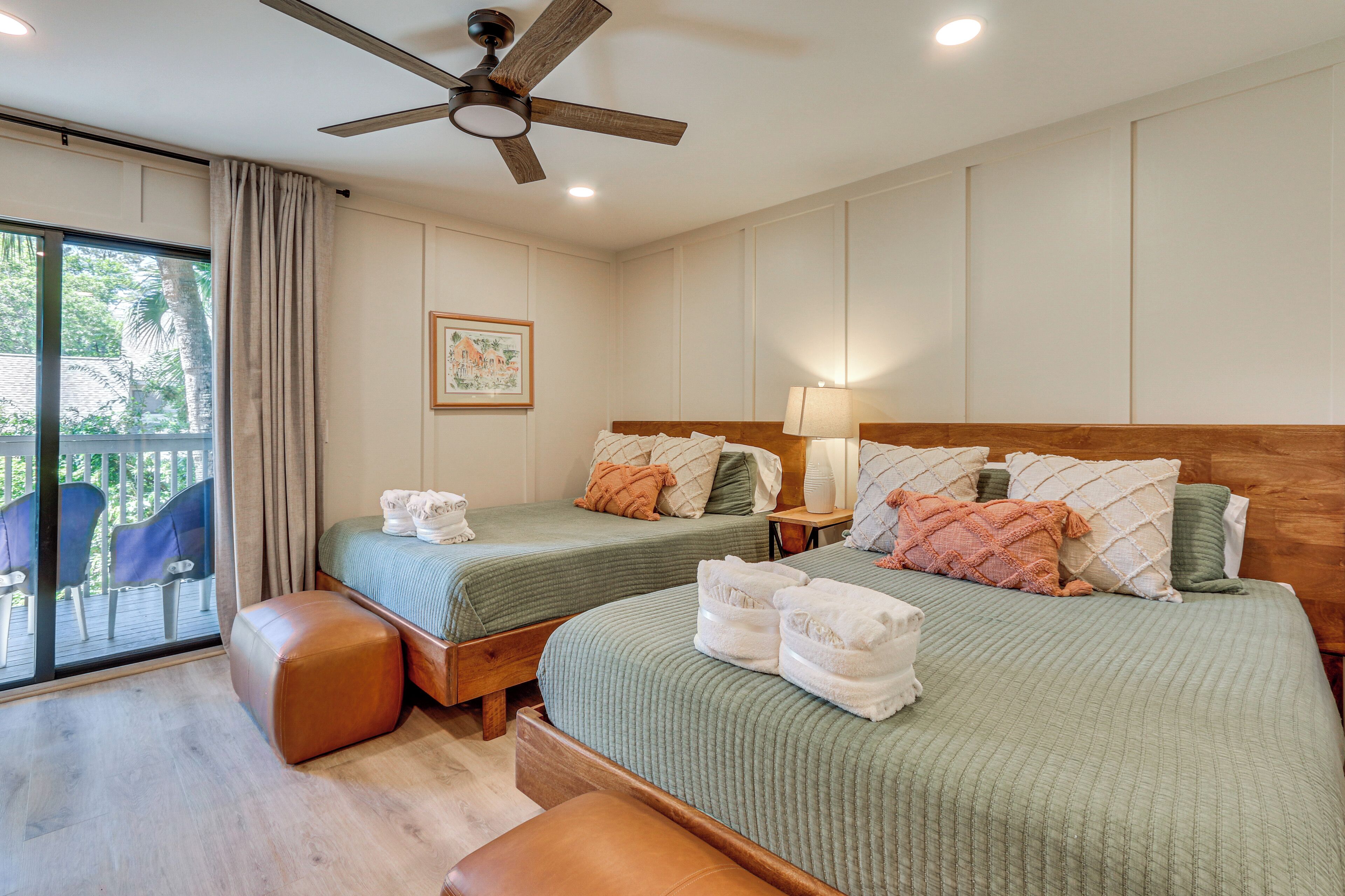 A hotel room with two beds, a ceiling fan, a large window with a view of palm trees, and a balcony with chairs.