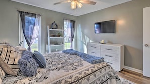 Une chambre à coucher comprenant un lit, un ventilateur de plafond, un téléviseur fixé au mur, une commode et une fenêtre avec des rideaux.