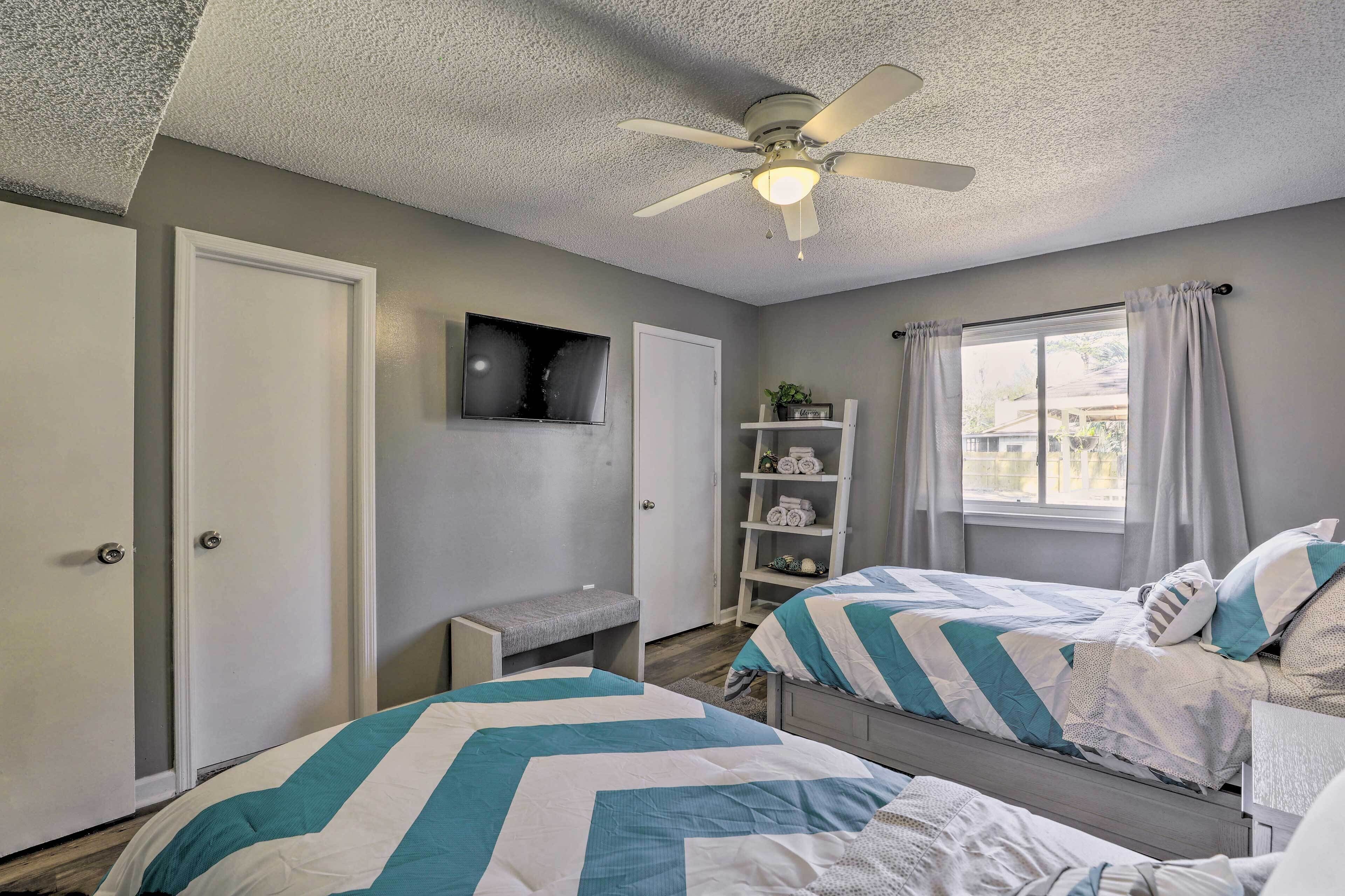 A bedroom with two beds, a ceiling fan, a TV, a shelf with towels, and a window with curtains.