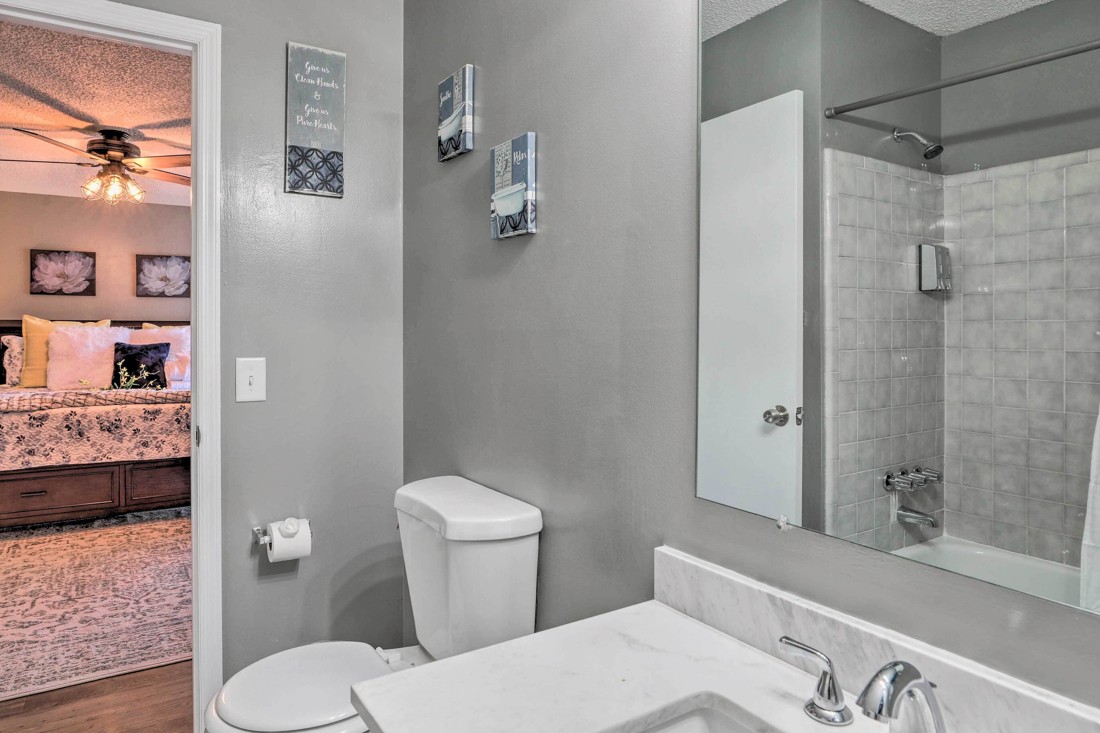 Une salle de bain avec des toilettes, un lavabo et une douche. Deux tableaux encadrés ornent le mur, et un ventilateur de plafond est installé au plafond.