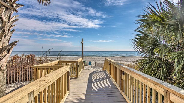 Ein hölzerner Steg führt zu einem Strand mit Palmen und blauem Himmel.