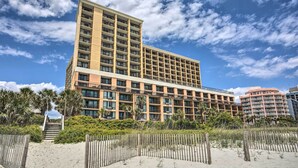 Ein mehrstöckiges Hotel mit flachem Dach und Balkonen, umgeben von einem Sandstrand und Palmen.