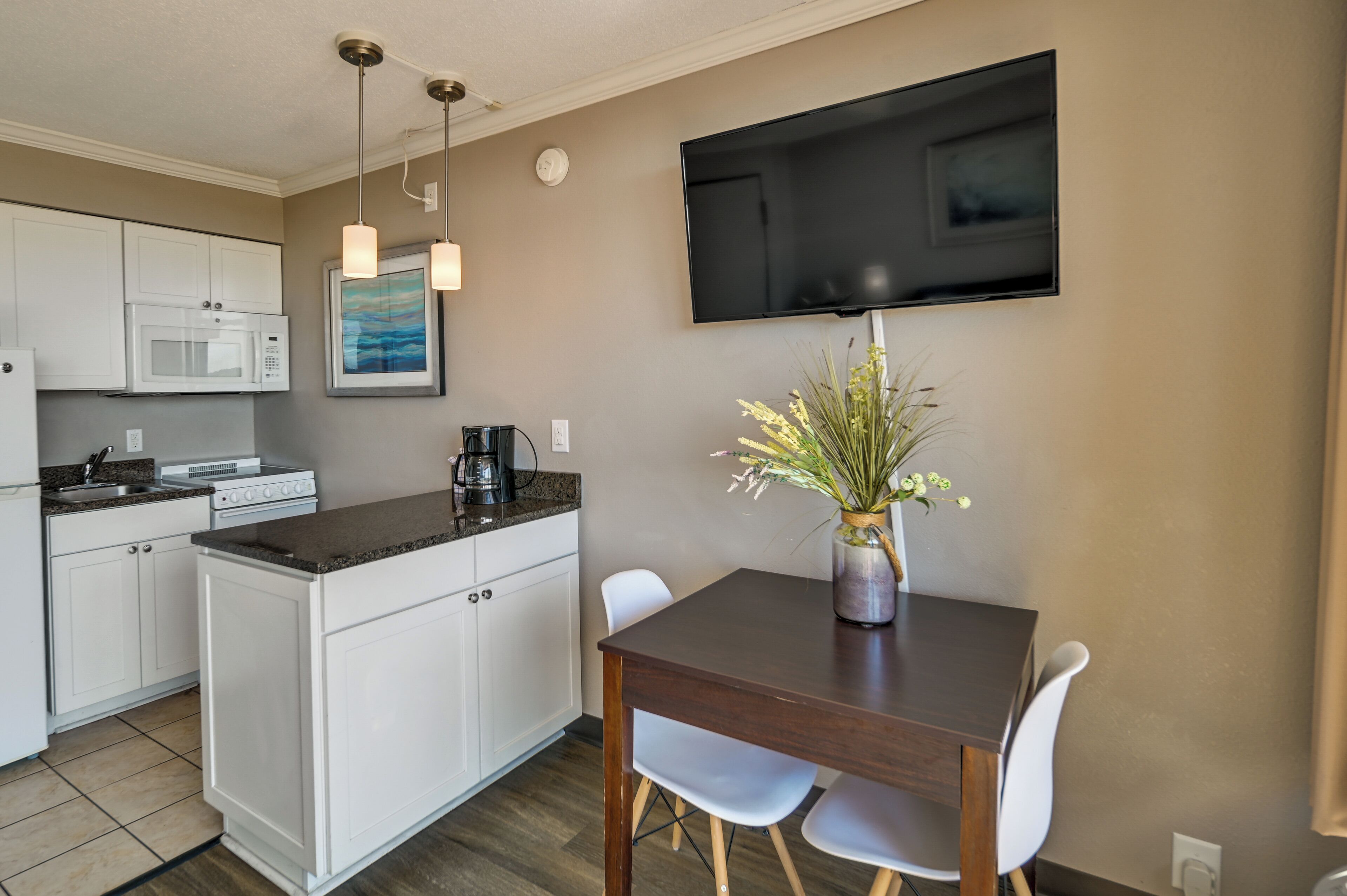 A compact kitchen with white cabinets, a microwave, and a coffee maker. A dining table with two chairs and a vase of flowers is in the dining area. A flat-screen TV is mounted on the wall.