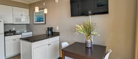 A compact kitchen with white cabinets, a microwave, and a coffee maker. A dining table with two chairs and a vase of flowers is in the dining area. A flat-screen TV is mounted on the wall.
