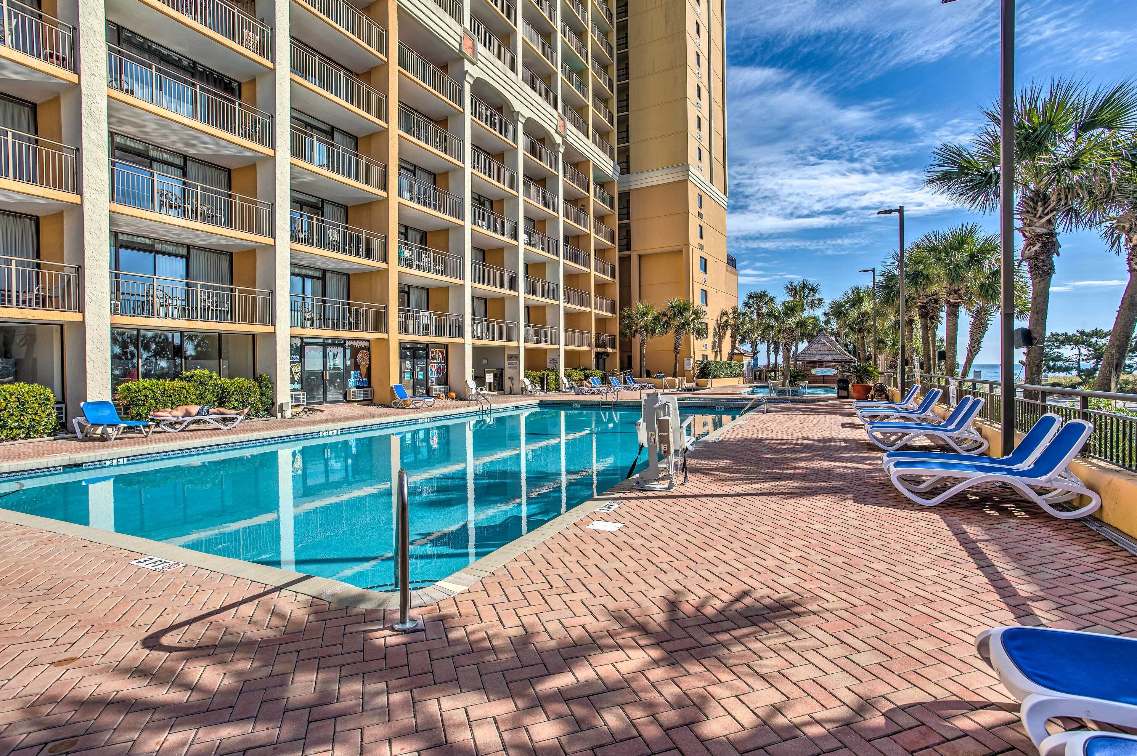 A hotel with a large outdoor swimming pool, sun loungers, and a palm tree-lined walkway.
