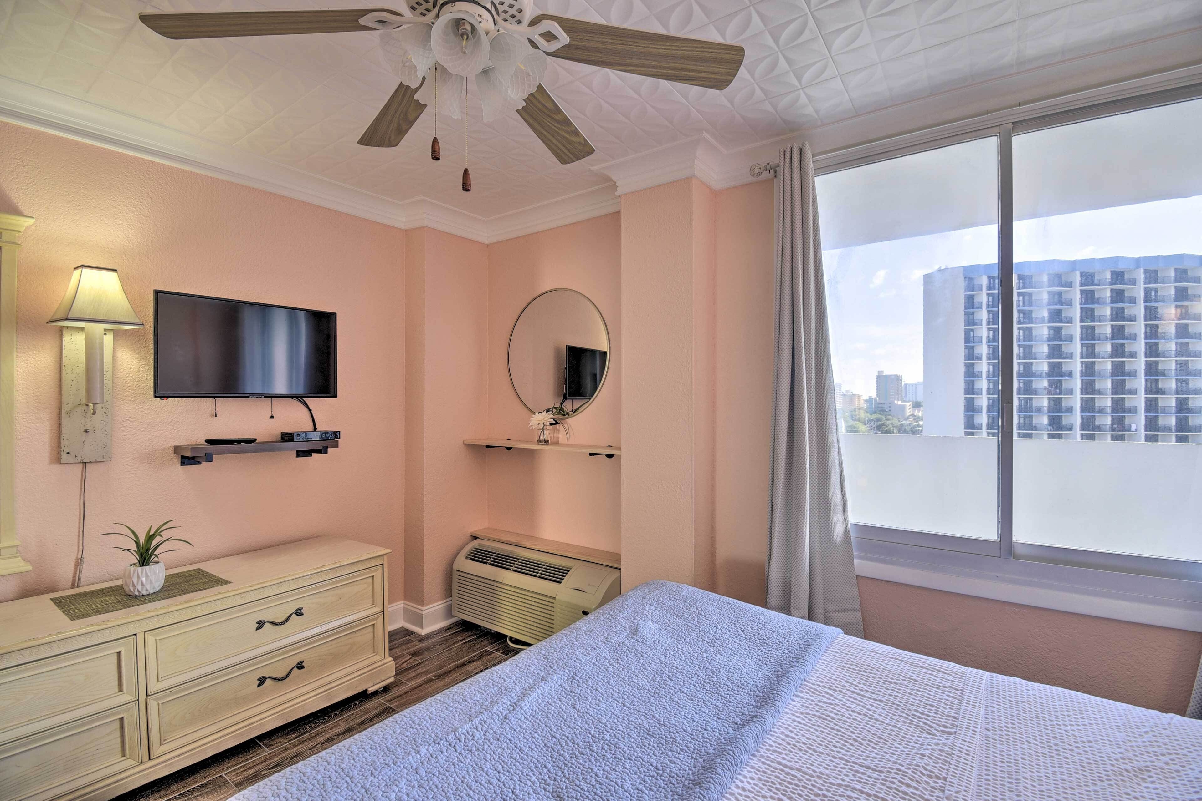 A bedroom with a bed, a ceiling fan, a TV, a mirror, a dresser, and a window with a city view.