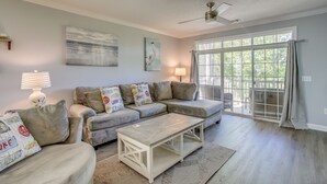 A living room with a sectional sofa, a coffee table, and a large window with a view of greenery. - North Myrtle Beach Condo w/ Golf Course Views! (North Myrtle Beach)