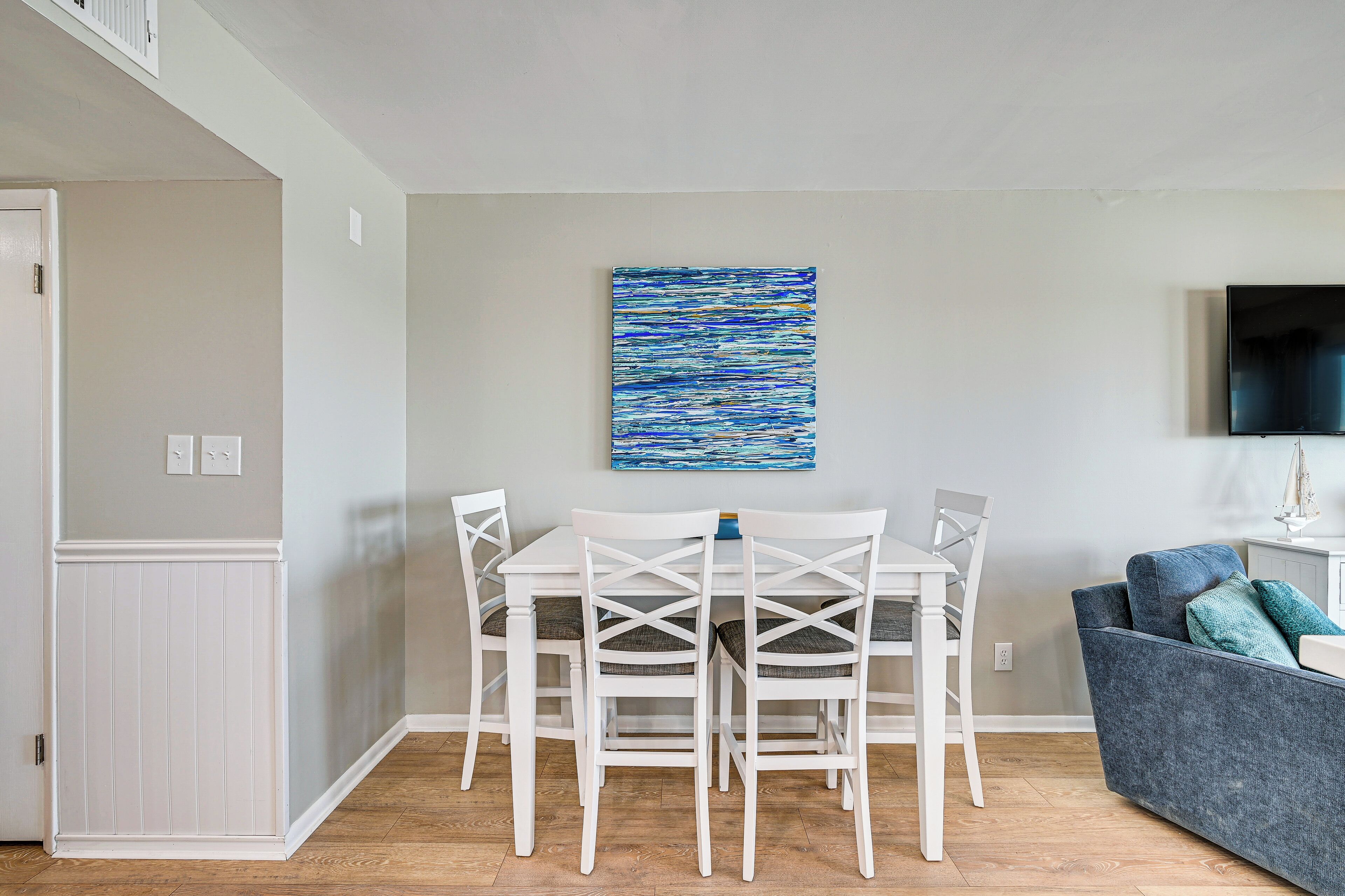 A dining area with a white table and chairs, a blue abstract painting on the wall, and a gray sofa with blue cushions.
