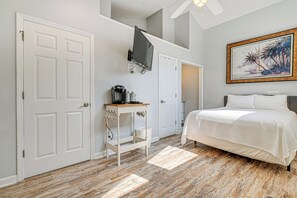 A bedroom with a bed, a coffee maker on a small table, a TV mounted on the wall, and a painting above the bed.