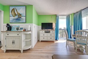 A room with a green wall, a painting of a sailboat, a television on a white dresser, and a balcony with blue curtains. - Oceanfront Myrtle Beach Rental w/ Pool! (Myrtle Beach)