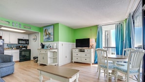 A living room with a dining area, kitchen, and a large window with curtains. - Oceanfront Myrtle Beach Rental w/ Pool! (Myrtle Beach)