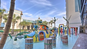 A water play area with a colorful submarine-themed structure, surrounded by palm trees and a building with a blue and white facade. - Oceanfront Myrtle Beach Rental w/ Pool! (Myrtle Beach)