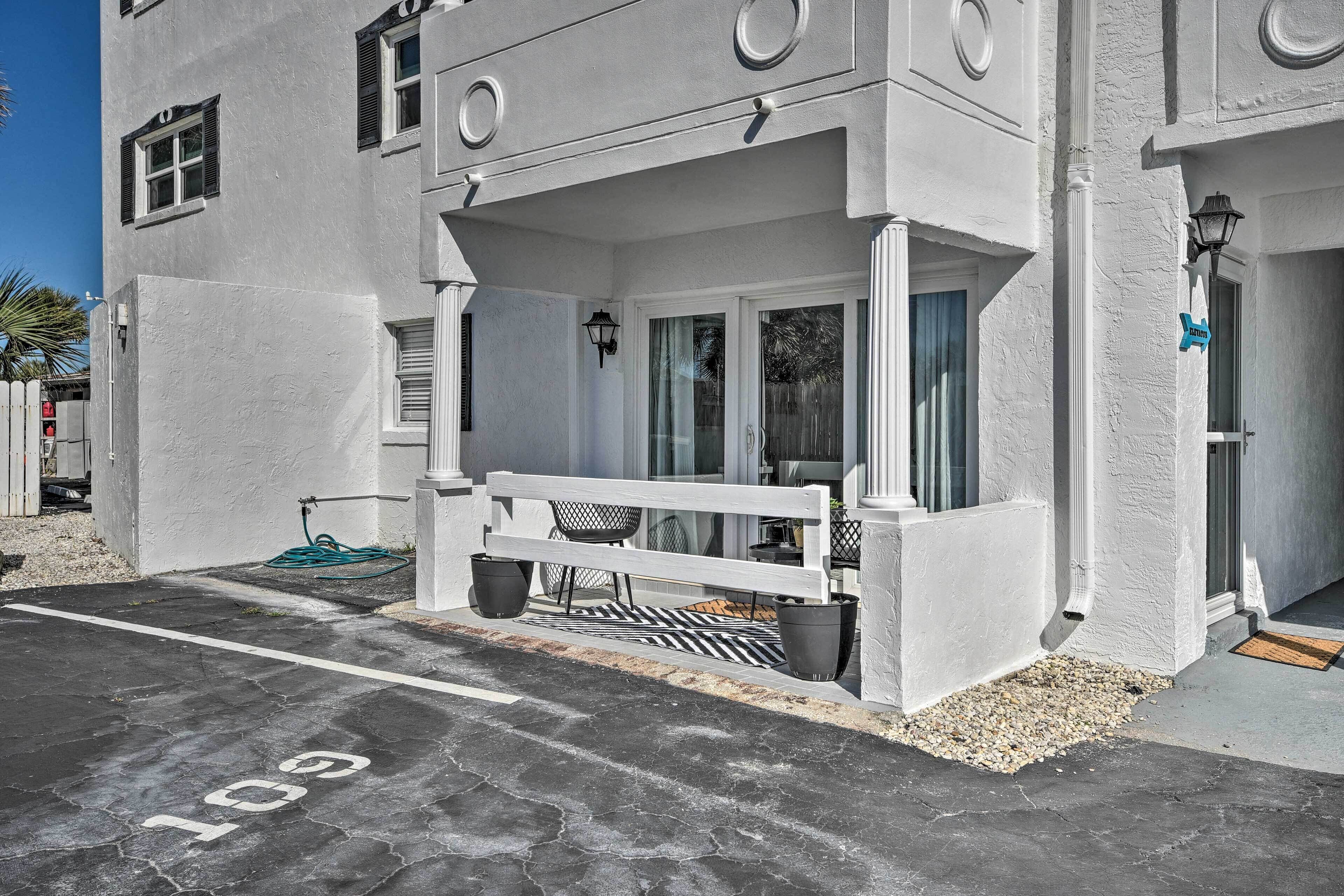 Ein weißes Gebäude mit überdachter Außenbereich-Sitzgelegenheit, einem ausgewiesenen Parkplatz 'G10' und einem kleinen Garten mit Bank und Blumentöpfen.