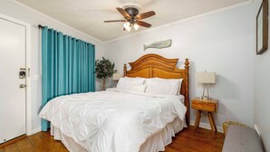 Une chambre à coucher bien rangée, avec un lit en bois, un ventilateur de plafond, une table de chevet avec une lampe et un panneau fixé au mur.