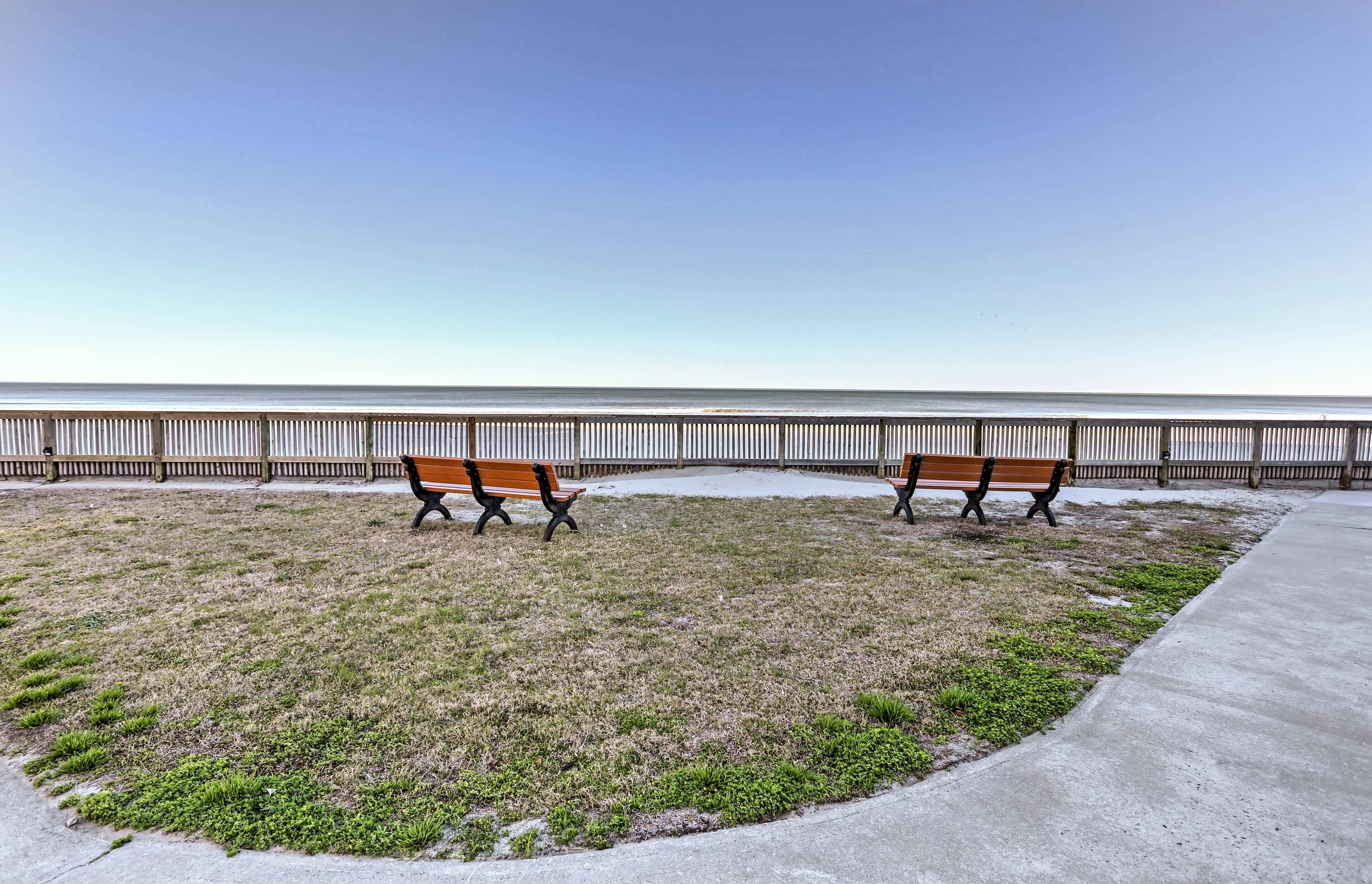 Une zone herbeuse avec deux bancs, disposés face à une clôture métallique qui donne sur une étendue d’eau.