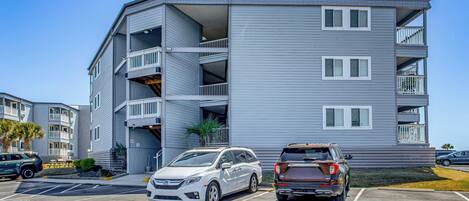 A multi-story building with balconies, parking spaces, and a clear sky.