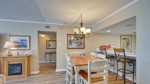 A dining area with a wooden table, white chairs, and a chandelier. There is a fireplace and a painting on the wall. - Oceanfront Myrtle Beach Condo w/ Outdoor Pool (Myrtle Beach)
