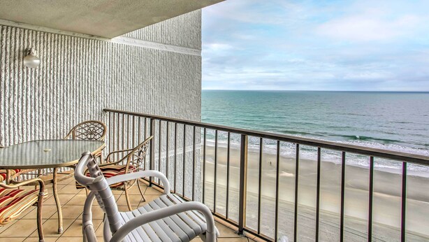 A balcony with a glass table and chairs, overlooking a beach with waves. - Oceanfront Myrtle Beach Condo w/ Outdoor Pool (Myrtle Beach)
