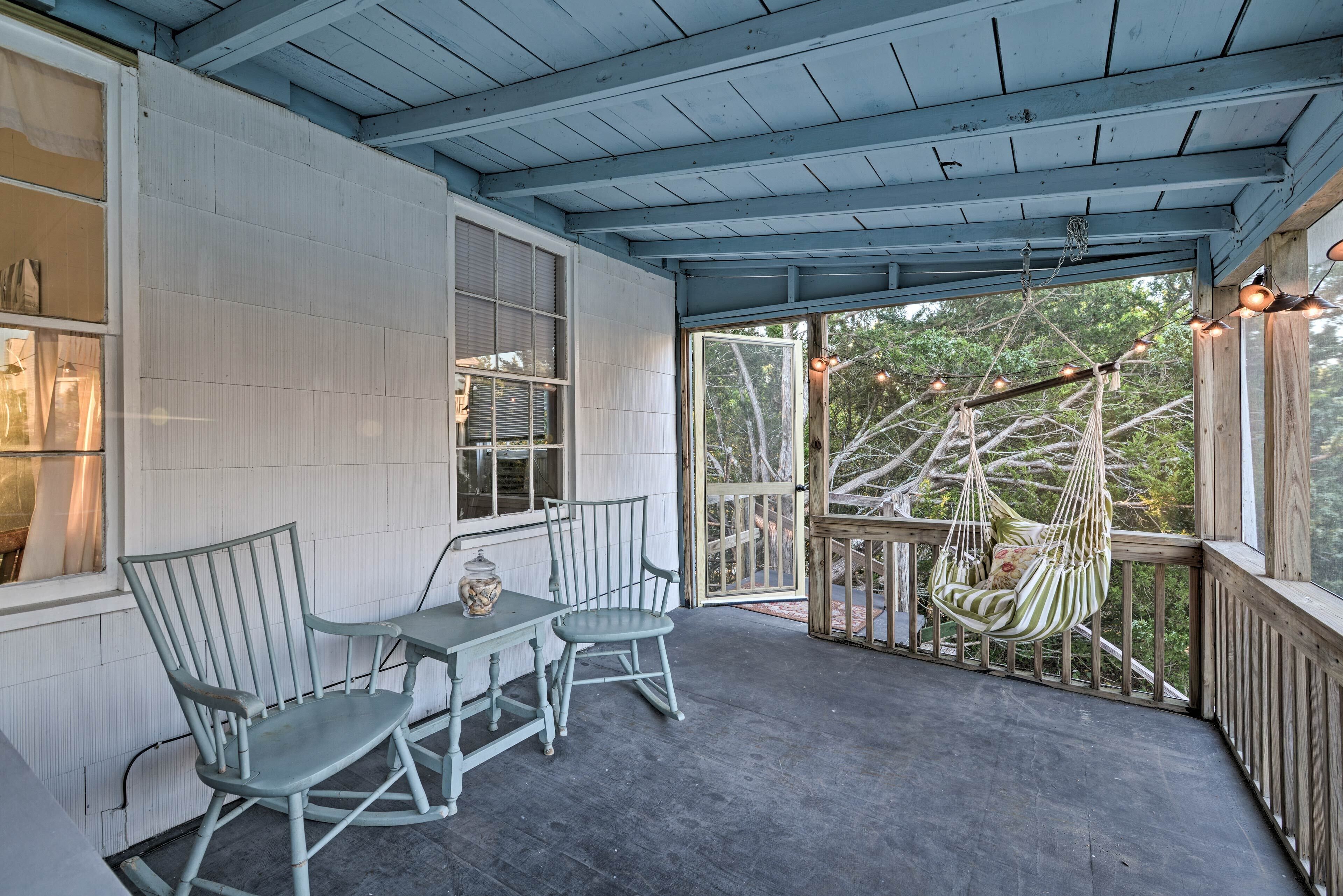 A covered porch with a table and chairs, a hanging chair, and a view of trees through the open door.