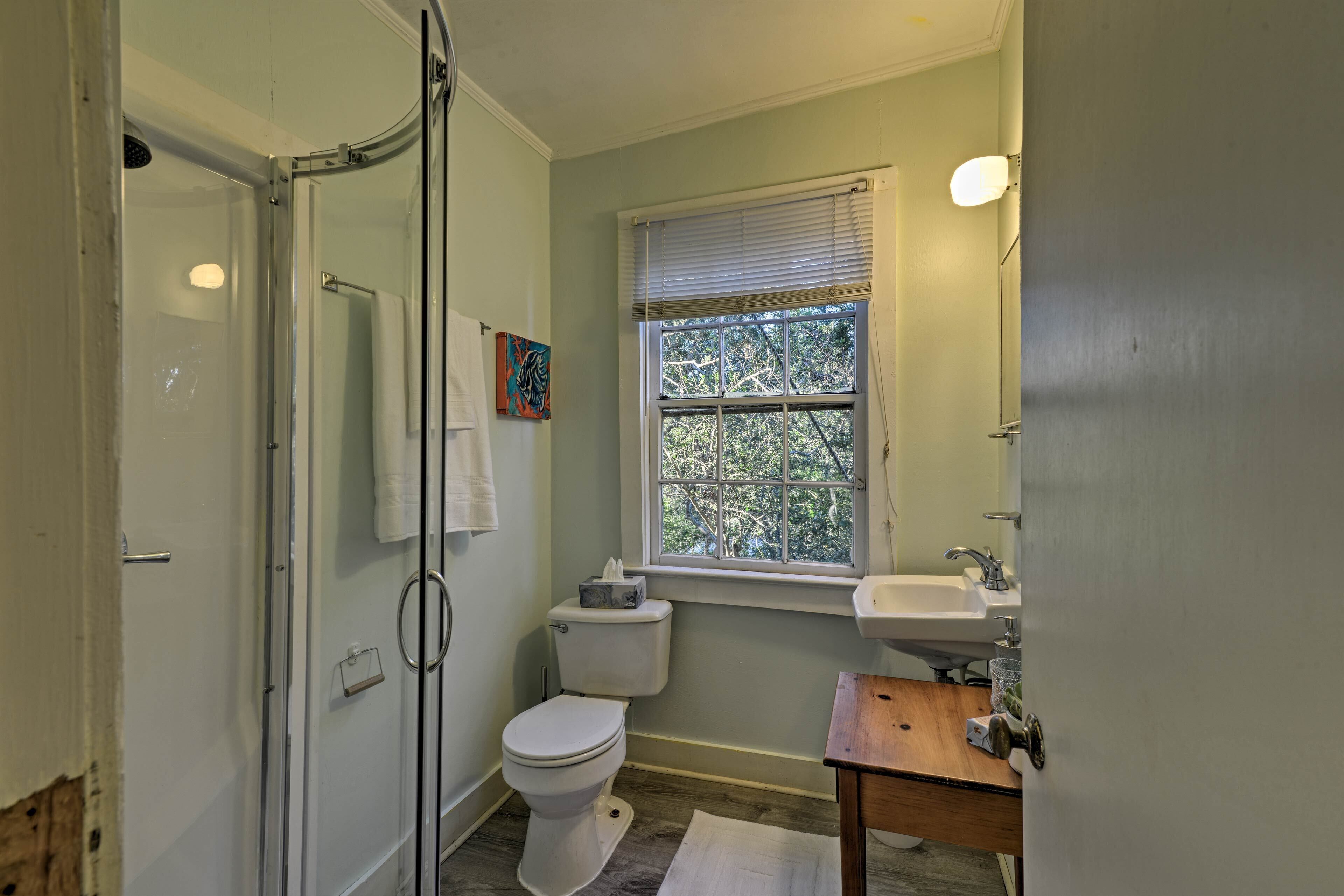A bathroom with a glass shower enclosure, a toilet, a sink, and a window with blinds.