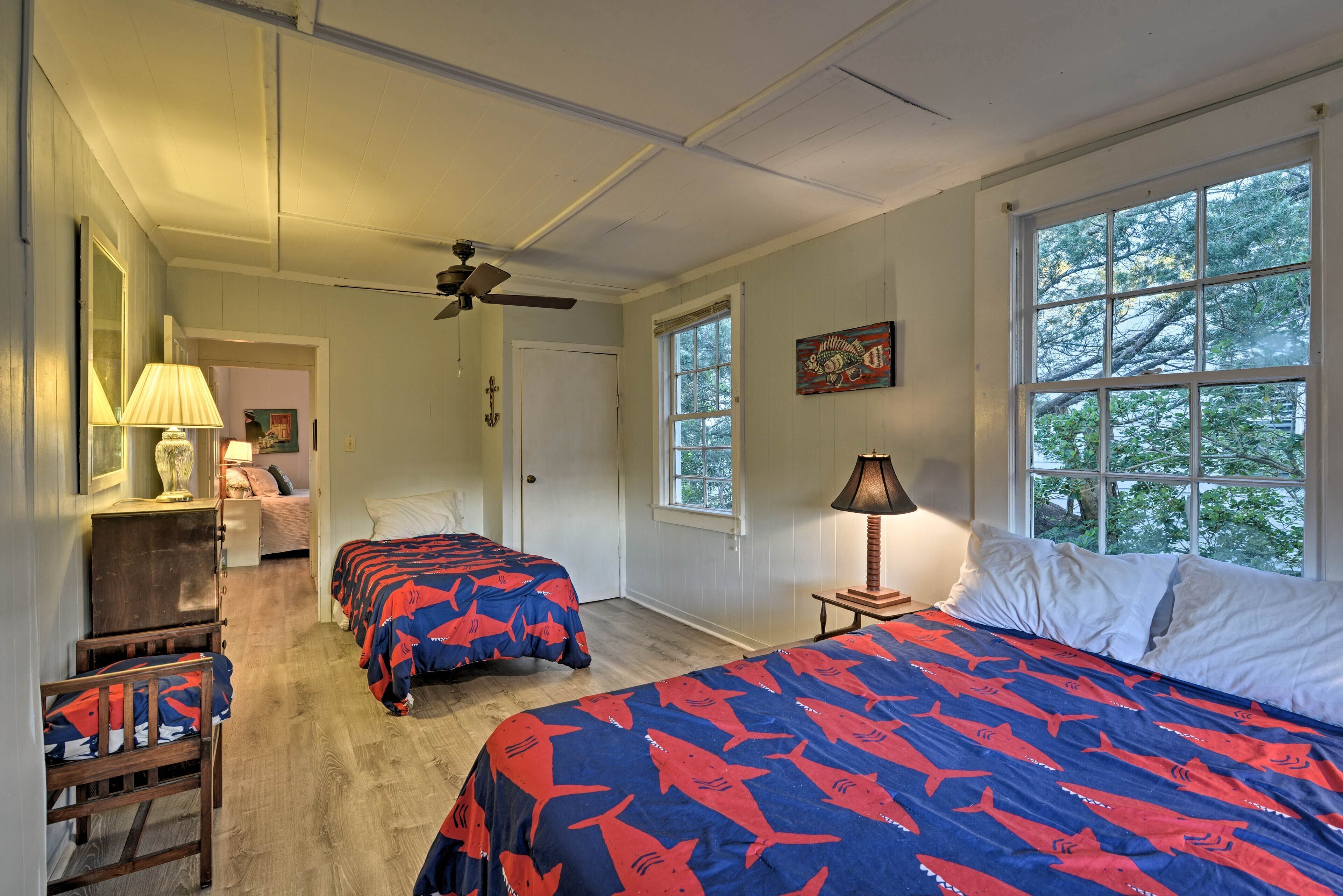 A bedroom with two beds, a ceiling fan, a window with a view of trees, and a door leading to another room.