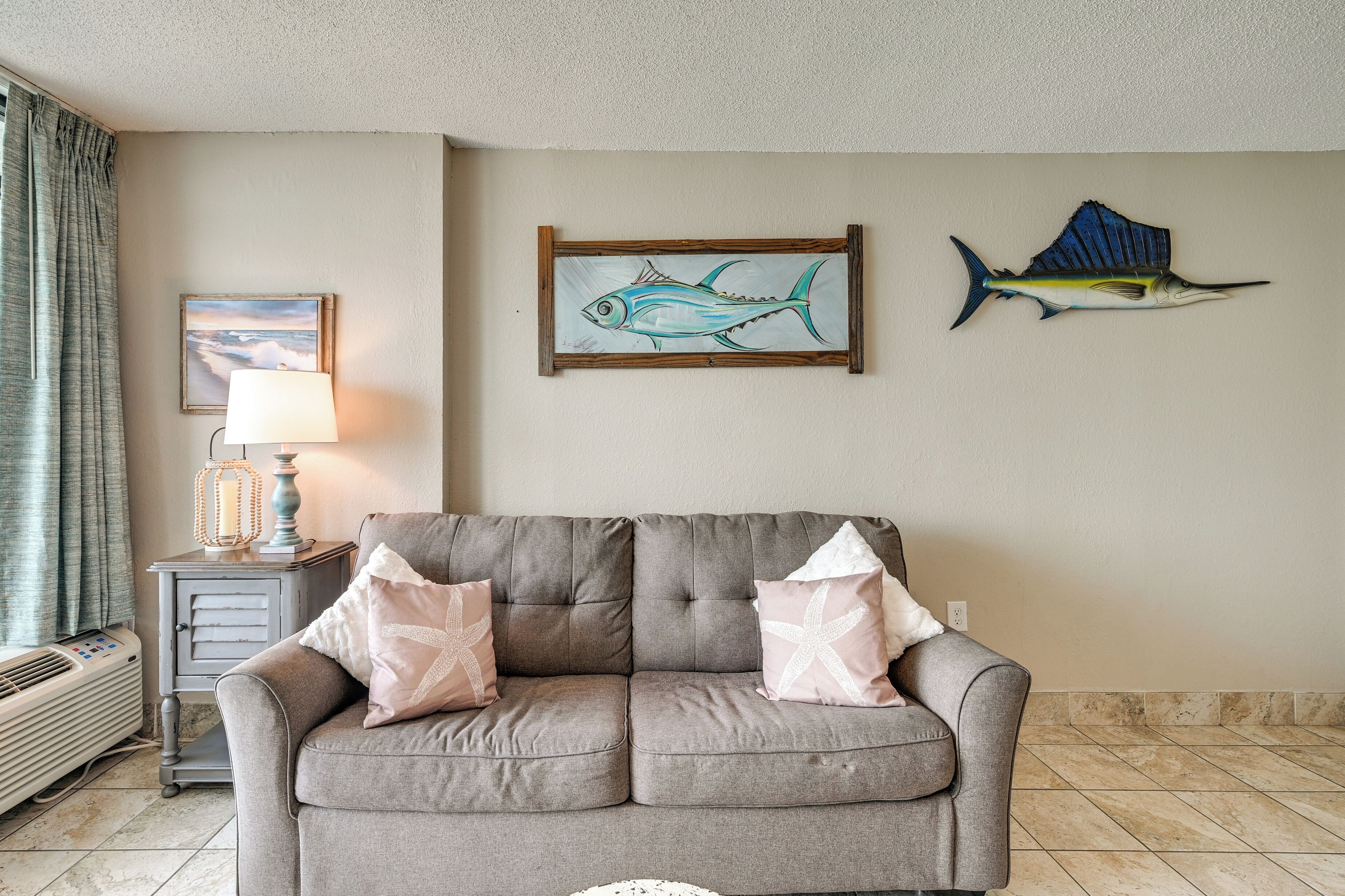 Una sala de estar con un sofá gris, dos almohadas con forma de estrella de mar, un cuadro de un pez enmarcado y una decoración de pez azul en la pared.